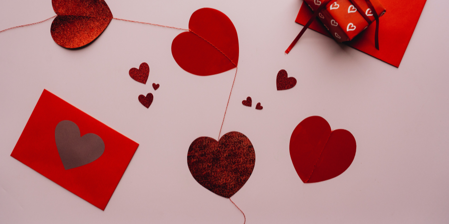 un fond rose pastel, sur lequel sont posés des cœurs en papier rouge, une enveloppe rouge avec un coeur dessus, ainsi qu'un cadeau rouge en haut à droite